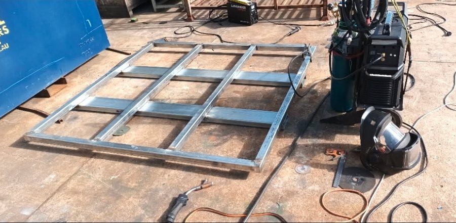 A metal frame with cross members lying flat on a concrete slab beside a MIG welder on a trolley, gas bottle, helmet and leads